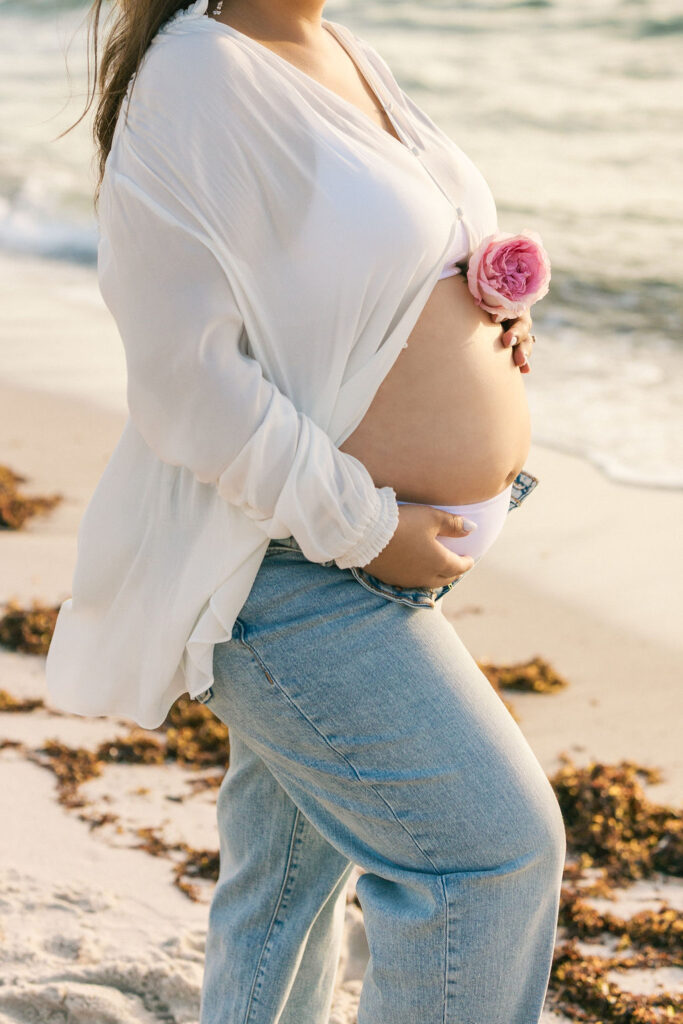 Maternity beach photography detail of baby bump with pink flower at sunrise in Miami.