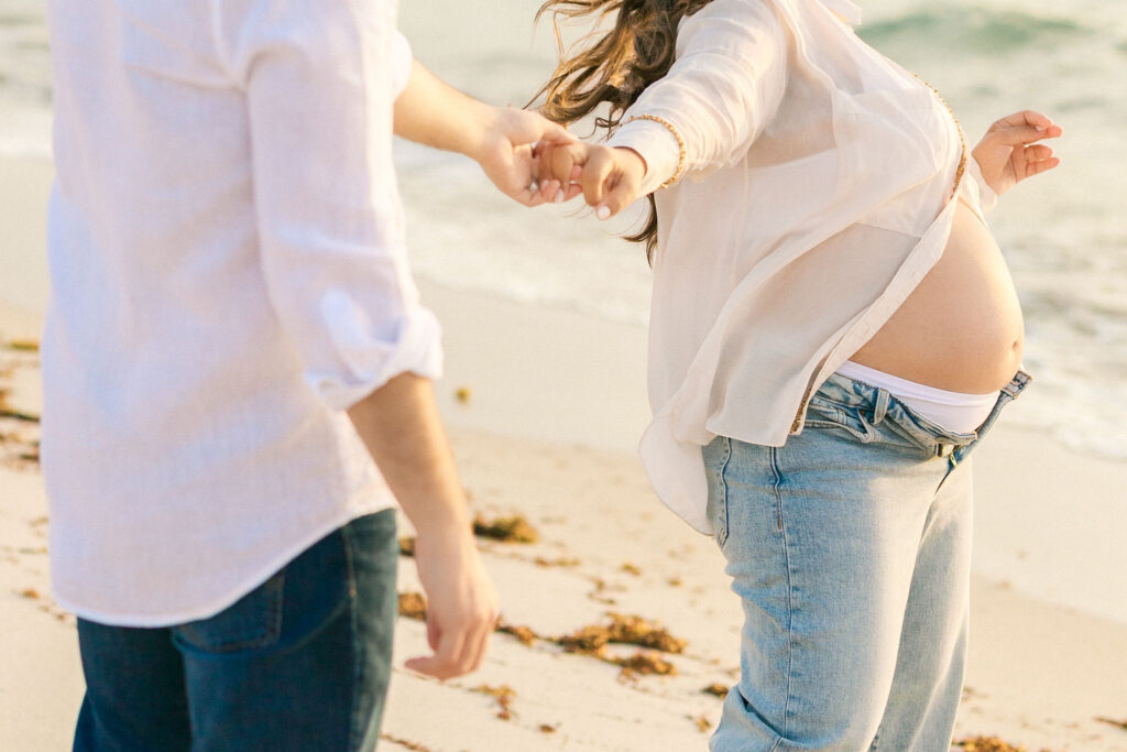 Playful maternity beach session with expecting couple holding hands at sunset in Miami.
