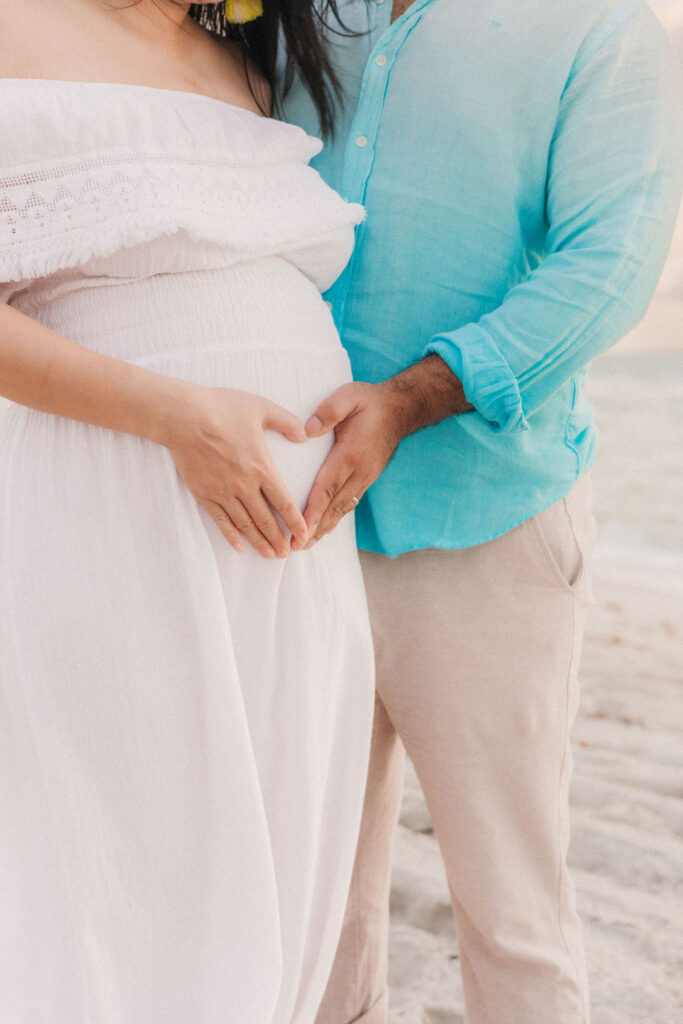 Parents forming a heart over baby bump during a maternity beach session.