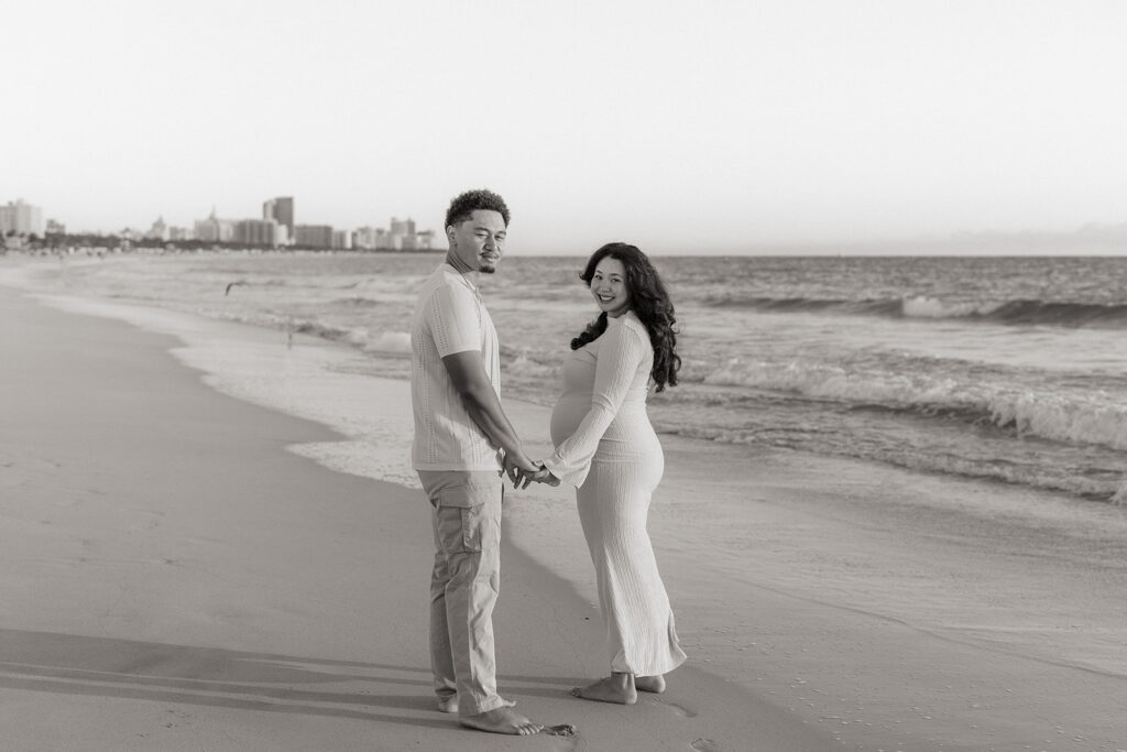 An expecting couple holding hands looks back while walking along the shoreline in Miami.